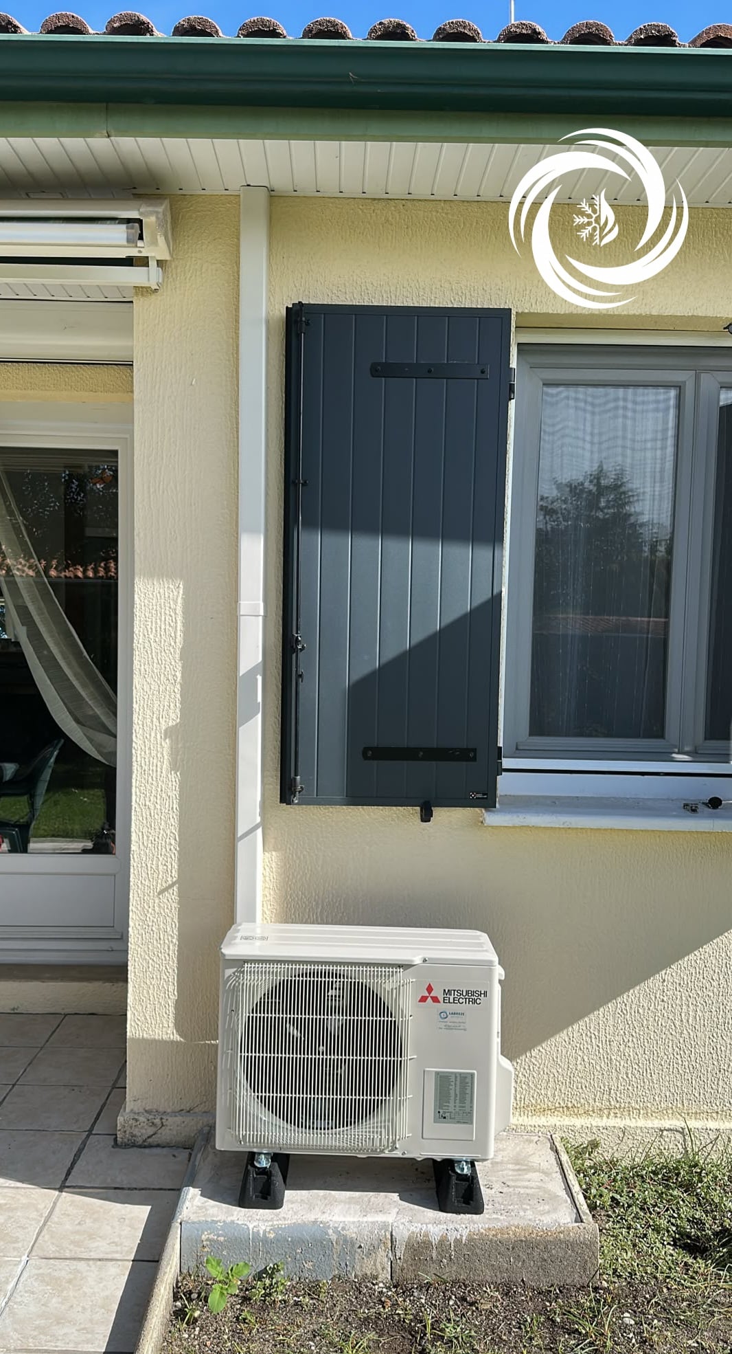Installation Climatisation Mitsubishi Electric sur Façade Unité extérieure Mitsubishi Electric installée sur un socle devant une façade jaune avec volet gris et fenêtre.